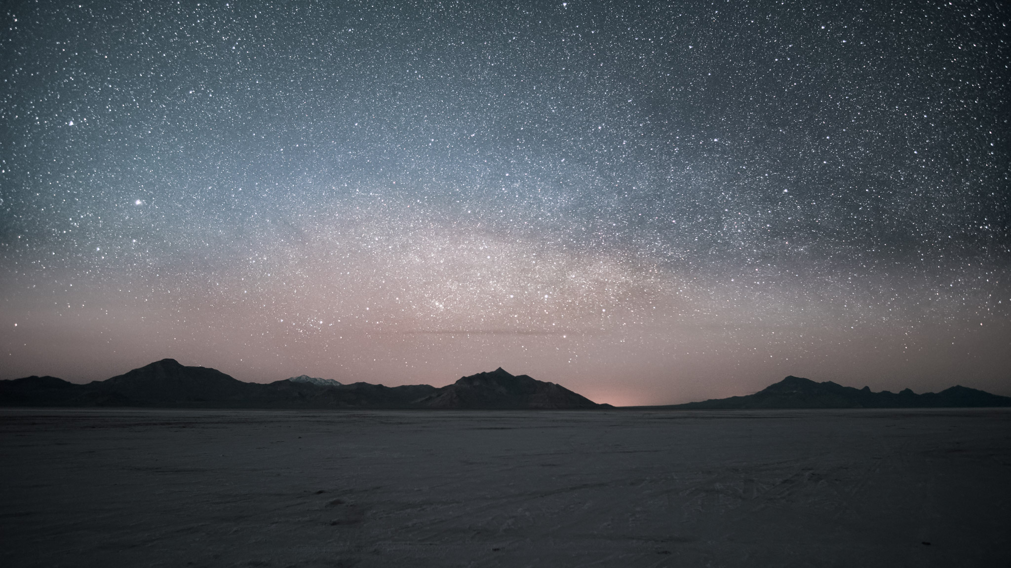 Over salt flats and distant black mountains the milky way spans, beneath it the sky is orangy from lingering sunset and above the sky is greeny blue